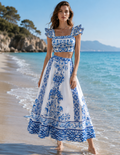Robe longue col V à bretelles volantées imprimé majolique bleu et blanc, galbe naturel au coucher du soleil sur la Méditerranée — collection ÉDITION SOLEIL 2026 | MITCH par AEF