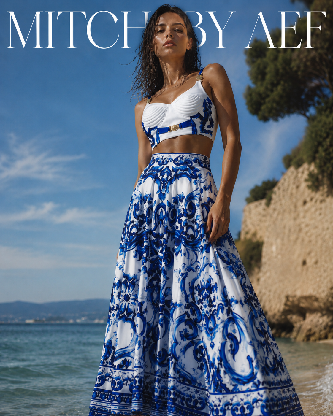 Mannequin professionnelle portant l’ensemble SANTORINI bleu et blanc, composé d’un bustier structuré et d’une jupe longue à imprimé baroque, photographiée de face sur une plage ensoleillée avec vue sur la mer.