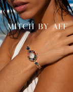 Montre bijou ÉCLAT dorée Arc-en-Ciel Joaillerie au poignet d'une femme à la plage, cadran à strass multicolores et bracelet fin en alliage doré, collection MITCH By AEF