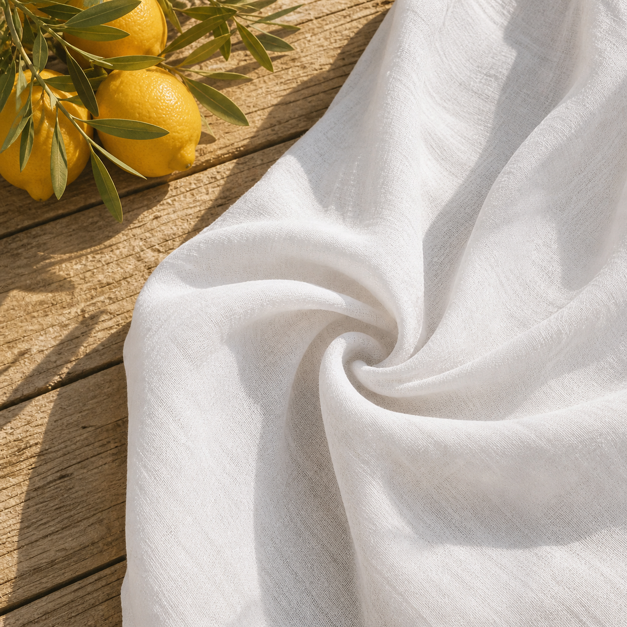 Détail texture tissu blanc fluide robe longue dos nu, posé sur bois flotté avec citrons frais et feuilles d'olivier, lumière naturelle soleil méditerranéen