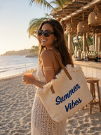 Sac cabas de plage en raphia naturel inscription Summer Vibes bleue anses cuir porté par une silhouette de mannequin en robe crochet blanche au bar de plage coucher de soleil cocktail à la main