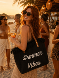 Sac cabas de plage en raphia noir inscription Summer Vibes blanche anses cuir franges noires porté par une silhouette de mannequin rousse au bar de plage coucher de soleil ambiance festive