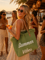 Sac cabas de plage en raphia vert sauge inscription Summer Vibes blanche anses cuir franges porté par une silhouette de mannequin blonde au bar de plage golden hour entourée d'amies avec cocktails